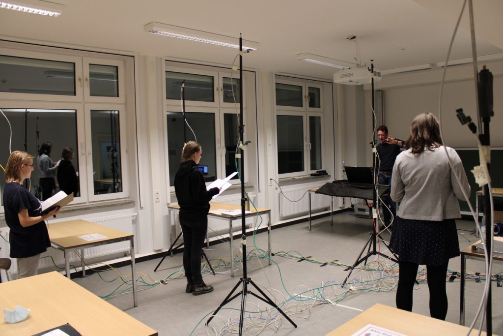 3 Sängerinnen und 1 Sänger (Chorleiter) in einem Seminarraum mit Stativen, Sensoren und Kabeln auf dem Boden während einer Chorprobe.