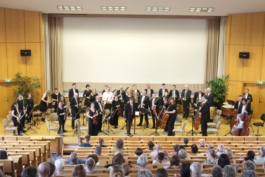 Das gesamte Orchester steht auf einer Bühne in einem holzgetäfelten Saal vor Zuschauern auf Sitzbänken.
