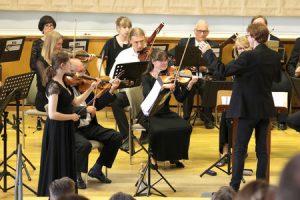 Musikerinnen und Musiker unterschiedlichen Alters mit Violinen, Bratschen, Fagott. Eine Violinen-Solistin stehend, die übrigen sitzend. Der Dirigent im dunklen Anzug hält die linke Hand erhoben.