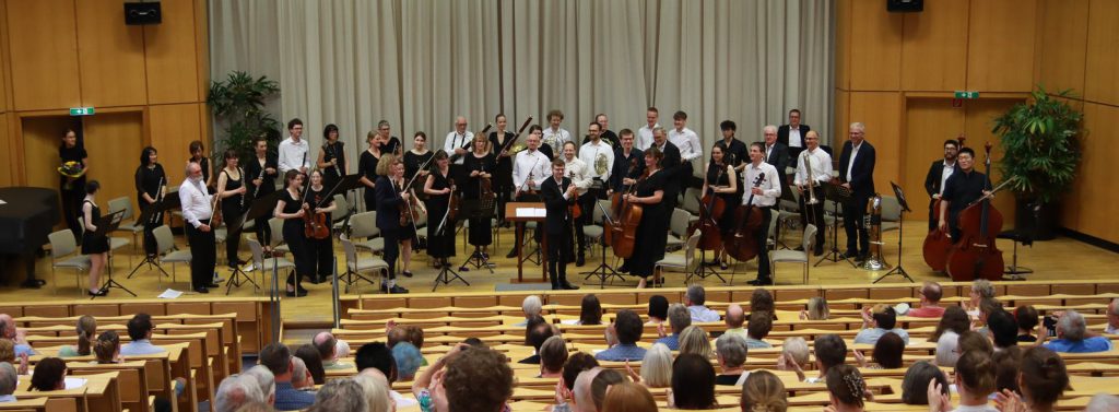 Ein Sinfonieorchester nach dem Konzert. Die Musiker und der Dirigent stehen, im Vordergrund des Fotos sieht man das Publikum in einem Hörsaal sitzen.
