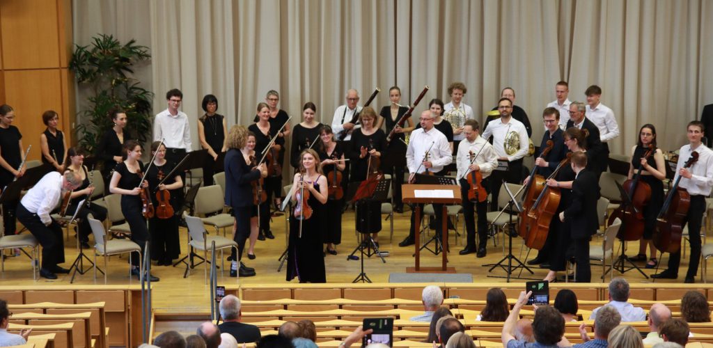 Ein Sinfonieorchester in schwarz-weißer Konzertkleidung und eine Violinsolistin im schwarzen Kleid stehen nach dem Konzert auf der Bühne in einem Hörsaal mit holzvertäfeltem Fußboden und Sitzreihen. Einzelne Personen im Publikum fotografieren mit Smartphones.